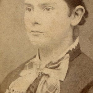 May include: Sepia-toned portrait of a person with dark hair pulled back. They are wearing a dark jacket with a decorative white scarf or cravat. The image is a vintage photograph, likely from the late 19th or early 20th century.