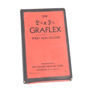 Puede incluir: Una caja rectangular de portapelícula plana Graflex con etiqueta roja y borde negro. El texto dice: ONE 2 1/4 x 3 1/4 GRAFLEX SHEET FILM HOLDER, TRADE MARK REG. U. S. PAT. OFF., MANUFACTURED BY THE FOLMER GRAFLEX CORP. ROCHESTER, N. Y., U. S. A.