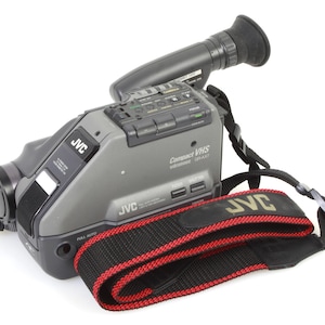 Könnte beinhalten: Ein grauer JVC Compact VHS-Camcorder mit einem schwarzen und roten Gurt. Der Camcorder verfügt über ein eingebautes Objektiv und eine Vielzahl von Knöpfen und Bedienelementen.