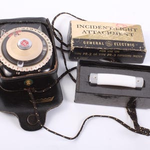 May include: A vintage General Electric light meter with a black leather case, a box labeled "Incident-Light Attachment," and a white cylindrical light attachment. The light meter has a gold-colored dial and a black strap.