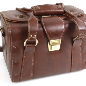 Puede incluir: Bolso de cámara de cuero marrón con asa superior y bandolera. El bolso tiene forma rectangular con un cierre y herrajes dorados. El bolso está hecho de un material de cuero texturizado.