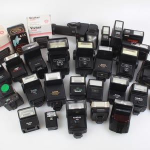 May include: A collection of black camera flashes, some with attached boxes of Vivitar 3500 film. The flashes vary in size and design, with some featuring silver or white reflectors. A few have visible brand names like Canon and Achiever.