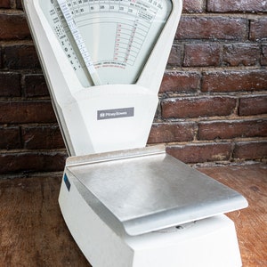 Puede incluir: Una báscula postal vintage Pitney Bowes blanca con una plataforma de pesaje plateada. La báscula tiene un dial circular con números del 0 al 20 y una tabla con tarifas postales para diferentes zonas.