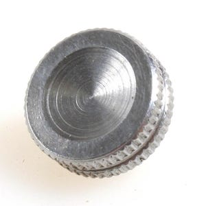May include: A close-up of a small, circular, silver-colored knob. The knob has a ridged edge and a flat, recessed center. The metal surface shows signs of wear and use, with subtle scratches and variations in tone. The background is plain white.