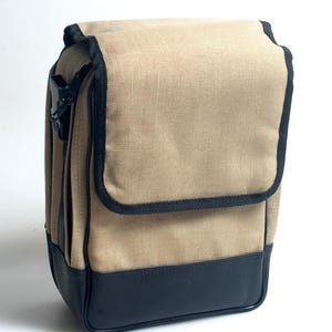 May include: A tan camera bag with a black leather base and trim. The bag has a flap closure and a black plastic buckle. The bag is rectangular and has a textured fabric appearance.