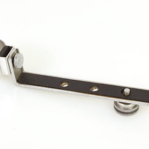 Puede incluir: Accesorio de cámara de metal plateado con una abrazadera circular, un brazo en ángulo recto y una base rectangular negra con tres agujeros. Se ve un botón roscado.