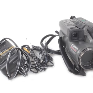 Puede incluir: Una videocámara VHS Panasonic gris con correa para el hombro, lente y visor negros. La videocámara va acompañada de un adaptador de corriente y cables negros. La lente tiene la inscripción "150x Digital".