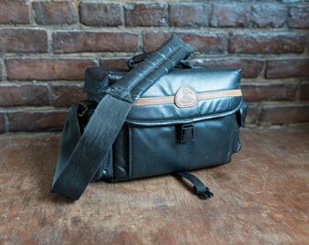 May include: A black camera bag with brown leather trim and a shoulder strap. The bag has a zipped pocket on the front and a buckle closure.