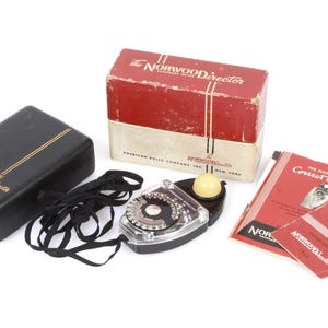 May include: A black case with gold lettering that reads "The Norwood Director" sits next to a black and silver light meter with a white dome. The light meter is attached to a black cord. A red box with white lettering that reads "The Norwood Director" sits behind the light meter. Two red pamphlets with white lettering that reads "The Manual for Correct Exposures" and "ASA Indexes" sit in front of the light meter.