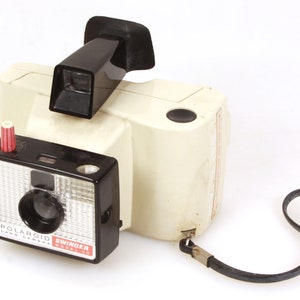 May include: A white Polaroid Swinger Model 20 Land Camera with a black viewfinder and a black strap. The camera has a red button on the side and a black lens.