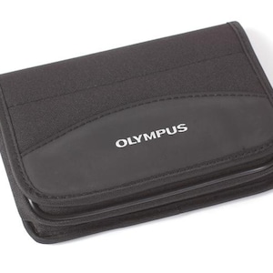 May include: Black Olympus camera case with a zip closure. The case is rectangular with rounded corners and a stitched design. The Olympus logo is printed in white on the front.