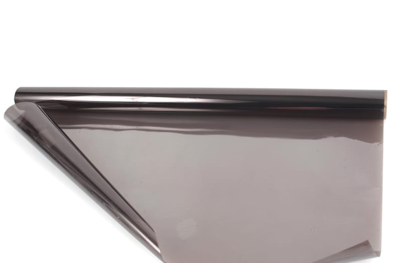 May include: A roll of tinted window film, partially unrolled, against a white background. The film is a dark gray color and appears to be reflective. The roll is cylindrical, and the unrolled portion lies flat.