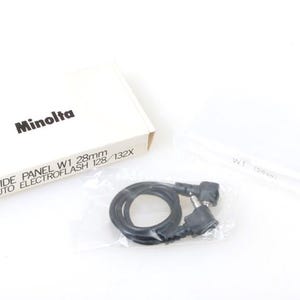May include: A white box with the text "Minolta Wide Panel W1 28mm Auto Electroflash 128/132X" on it. The box is next to a clear plastic bag containing a black cable with two connectors.