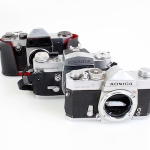 May include: A collection of vintage cameras, including a Konica Autoreflex, a Miranda, and a Sensorex. The cameras are black and silver, with leather accents and various dials and lenses. The Konica camera has a large lens opening.