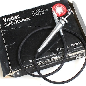 Puede incluir: Una caja negra con texto blanco que dice "Vivitar Cable Release" y "For Vivitar Quick Release Pistol Grip". Un disparador de cable plateado con un cable negro está encima de la caja.