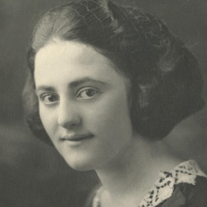 May include: A black and white vintage portrait of a person with dark hair styled in a bob with a fine hairnet. The person wears a dark garment featuring a white lace collar with an intricate openwork pattern along the neckline and shoulders.