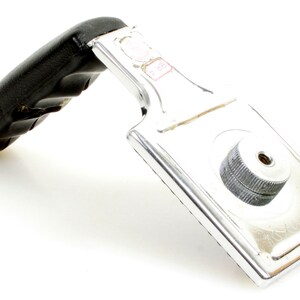 May include: A chrome-plated metal handle with a black, padded grip. The handle has a rectangular base with a circular, ridged knob. The grip shows signs of wear. The item is likely a replacement part.