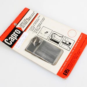 May include: A silver camera clip with a screw for attaching to a camera strap or belt. The packaging says "Capro Camera Clip for 110 System and other camera makes. Fits Kodak models 30 & 40. Sturdy, stainless steel clip with retaining screw. Holds camera securely to belt, camera strap or pocket. Capro Photo Products - The Ultimate in Quality. EPI Ehrenreich Photo Optical Industries, Inc. Garden City, New York."