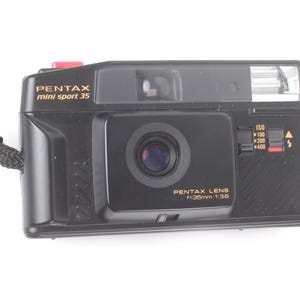 May include: Black Pentax mini sport 35 camera with a black strap. The camera has a lens with the text "PENTAX LENS f=35mm 1:3.8". The camera has a red button and a flash.