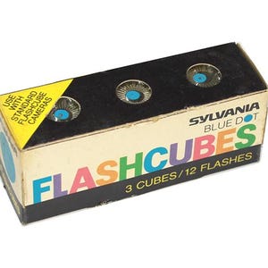 May include: A box of three Sylvania Blue Dot Flashcubes. The box is black with the words "FLASHCUBES" in rainbow colors. The box says "3 CUBES/12 FLASHES" and "USE WITH STANDARD FLASHCUBE CAMERAS".