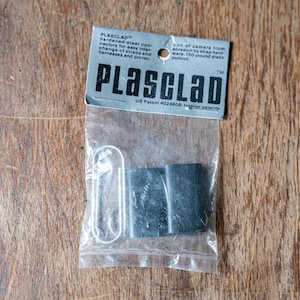 以下が含まれることがあります： 銀色の金属クリップと2つの黒い長方形のコネクタが入った透明なビニール袋。袋には「PLASCLAD」の文字と製品情報が記載された灰色のラベルが付いています。木製の表面に置かれたアイテムです。