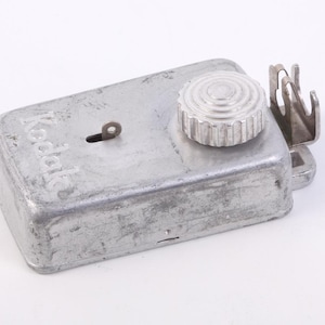 Op de afbeelding: Vintage Kodak film ontwikkel tank. De zilverkleurige metalen tank heeft een sleutelgat en een geribbelde knop. Het Kodak-logo is zichtbaar. Een metalen stuk is aan de zijkant van de tank bevestigd.