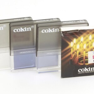 May include: Four grey plastic boxes with the word "cokin" printed on them. The boxes are labelled "Fog 2 A", "Graduated 81 A", "Diffuser 1 A", and "Chromopfilter 52 M". The boxes are arranged in a row from left to right. A box with the word "cokin" printed on it is in the foreground. The box has a picture of candles on it. The text "Creative Filter System" is printed on the box.
