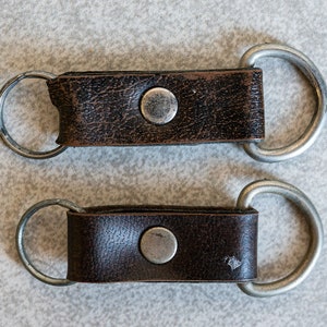 以下が含まれることがあります： 銀色の金属製のリングとボタンが付いた、茶色の革製のキーホルダーが2つ。