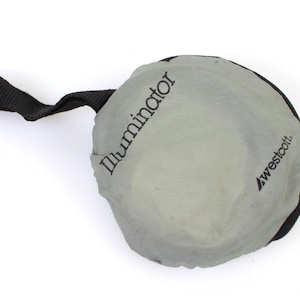 Könnte beinhalten: Ein runder, grauer Stoffreflektor mit einem schwarzen Riemen und dem aufgedruckten Text "Illuminator" und "Westcott".