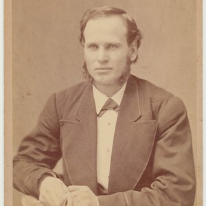 Pode incluir: Um retrato em preto e branco de um homem vestindo um terno escuro e gravata borboleta. Ele tem barba e bigode e está olhando diretamente para a câmera.