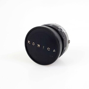 May include: A black Konica camera lens cap with gold lettering. The cap is round and has a ridged edge. The lens cap is on a white background.