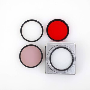 Puede incluir: Un conjunto de filtros de lente de cámara, que incluye un filtro rojo, rosa y transparente, junto con dos anillos negros vacíos. Un filtro está en una caja plateada. Los filtros son circulares.