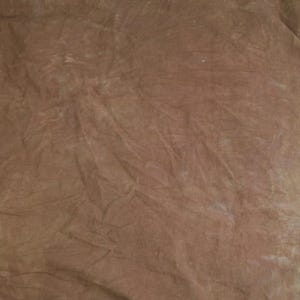 May include: A piece of fabric with a mottled, uneven brown dye pattern. The material features a series of soft, diagonal creases and folds across the surface, creating a textured appearance with varying shades of light and dark brown.