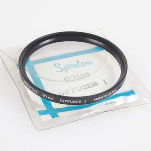 Puede incluir: Un filtro de lente de cámara Spiratone 67 mm Diffuser 1 negro en su embalaje original. El filtro está fabricado en Japón y se encuentra en una funda de plástico transparente con texto azul y blanco. El filtro es circular con un borde negro.
