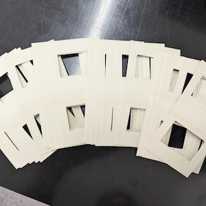 May include: A collection of off-white picture mats arranged in a fan shape. Each mat has a rectangular opening in the center and a beveled edge. The mats are stacked on a dark surface, creating a contrast.