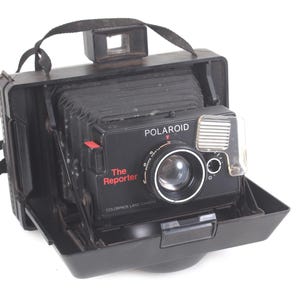 May include: Black Polaroid "The Reporter" camera with a lens, flash, and viewfinder. The camera is in an open black case with a black strap. The camera has red accents and the word "Polaroid" is visible.