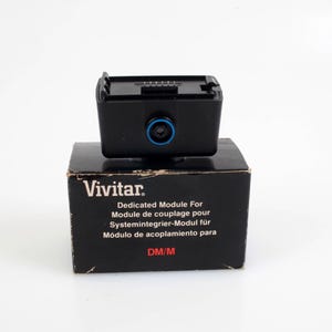 May include: A black Vivitar Dedicated Module For camera accessory is placed on its original box. The module has a blue accent around the lens and is designed for system integration. The box features text in multiple languages.
