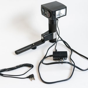 Puede incluir: Un flash negro con un brazo giratorio y un cable en espiral. El flash tiene un cable de alimentación con un enchufe británico.