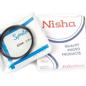 May include: A black 49mm Spiratone Diffuser filter and a Nisha Quality Photo Products box. The filter is in a clear plastic package with blue accents. The Nisha box is white with red and blue text.