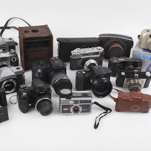 May include: A collection of vintage cameras, including SLR and point-and-shoot models, in various sizes and colors. Some cameras have lenses attached, and others are in cases or boxes. The cameras are arranged on a white surface.