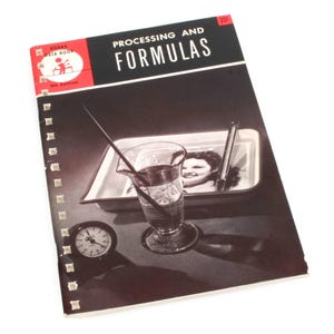 Peut inclure: Une photo en noir et blanc d'un visage de femme dans un bac de développement. Le bac est sur une surface sombre avec un bécher en verre et une baguette d'agitation. La couverture du livre indique "Traitement et formules" avec les mots "Kodak Data Book" dans un encadré rouge en haut à gauche.