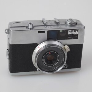 Könnte beinhalten: Eine Vintage Petri 7 Half Kamera mit silbernem Oberteil und schwarzem Gehäuse. Die Kamera hat ein markantes Objektiv mit silbernem Ring und der Text "PETRI 7 HALF" ist sichtbar. Ein klassisches Beispiel für Fotoausrüstung aus der Mitte des Jahrhunderts.