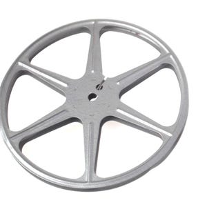 Puede incluir: Una bobina de película plateada con seis radios que irradian desde un cubo central. La bobina es de metal y tiene forma circular, diseñada para contener película. Los radios están espaciados uniformemente.
