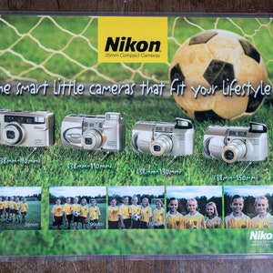 May include: A promotional poster for Nikon 35mm compact cameras. The poster features a soccer ball in front of a green grassy field with a white net. The text on the poster reads "The smart little cameras that fit your lifestyle". Four Nikon cameras are shown with their zoom ranges listed below each camera.