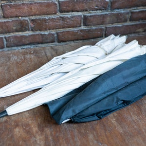 Könnte beinhalten: Zwei weiße Regenschirme und ein schwarzer Regenschirm, zusammengefaltet und auf einer Holzoberfläche gestapelt.