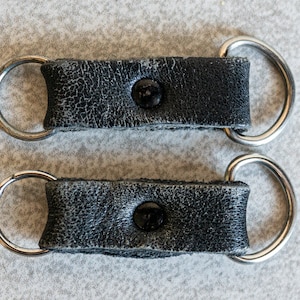 以下が含まれることがあります： 銀色の金属製のリングと黒いボタンが付いた黒い革のストラップが2本。ストラップはキーホルダーやバッグストラップとして使用するために設計されています。