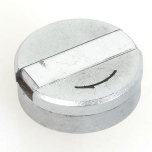Puede incluir: Pequeño objeto metálico redondo de color plateado, con una inserción rectangular blanca y una línea negra curva. El objeto tiene una forma circular con una parte superior plana y un borde ligeramente elevado.