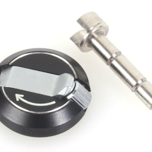 Puede incluir: Un componente negro y plateado con un diseño circular y una palanca plateada. Un tornillo cilíndrico plateado está posicionado junto al componente. El componente tiene una flecha blanca que indica la dirección.