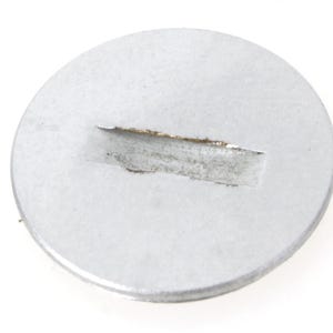 Puede incluir: Un objeto metálico circular de color plateado con una hendidura rectangular. El objeto parece ser un disco pequeño y plano, posiblemente un componente o parte de un mecanismo más grande. La superficie muestra signos de desgaste.