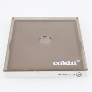 Puede incluir: Un portafiltros rectangular de plástico teñido de marrón con la marca "cokin" en blanco. El texto "DIFFUSER 2 P (084)" está impreso en la parte inferior. El portafiltros tiene una abertura circular transparente en el centro.
